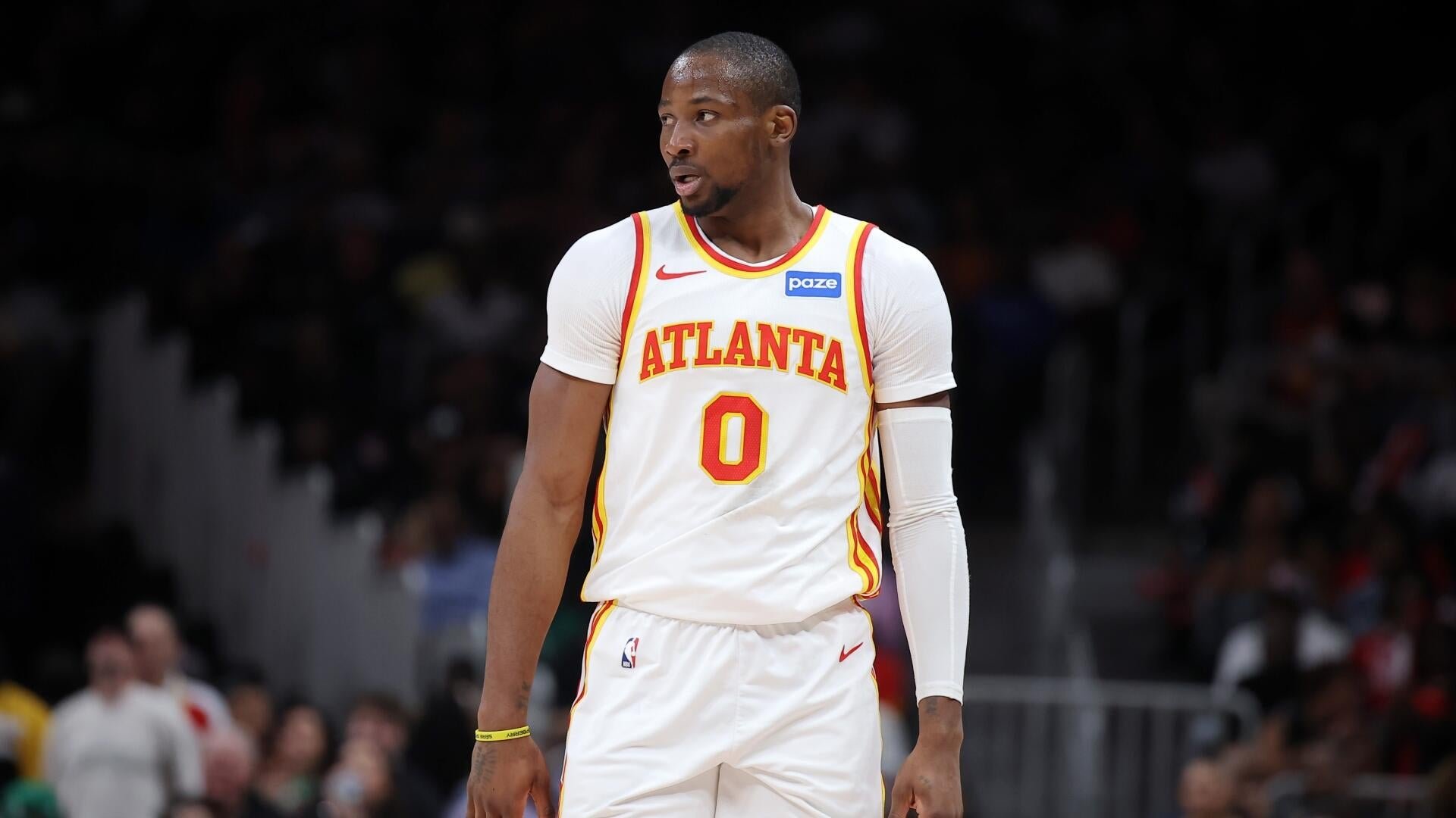 An Atlanta basketball player wearing jersey number 0 and a white arm sleeve stands on the court during a game, with a blurred crowd visible in the background.