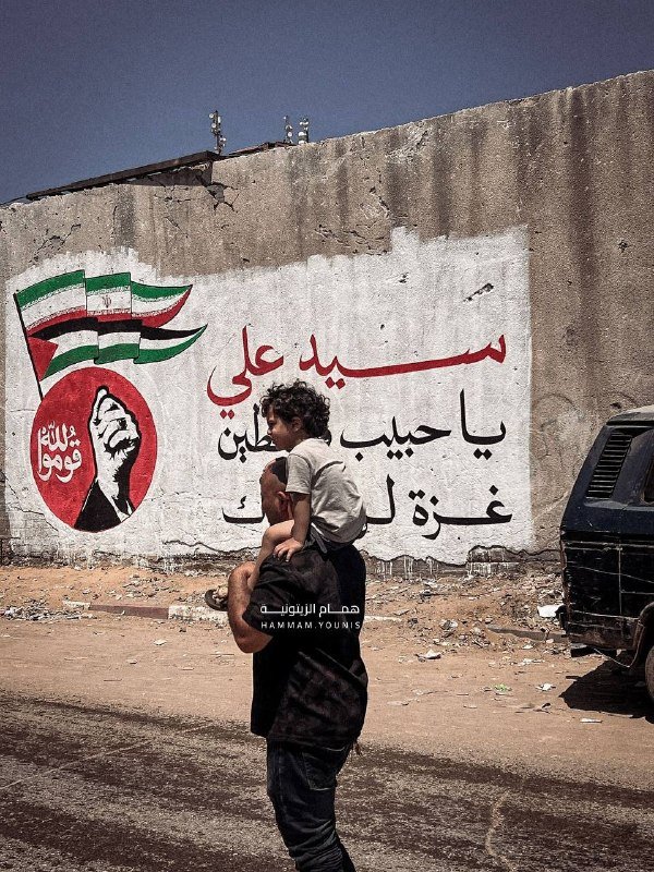 A man carries a young child on his shoulders along a dirt road past a weathered wall painted with Arabic text, a raised fist, and Syrian flags.