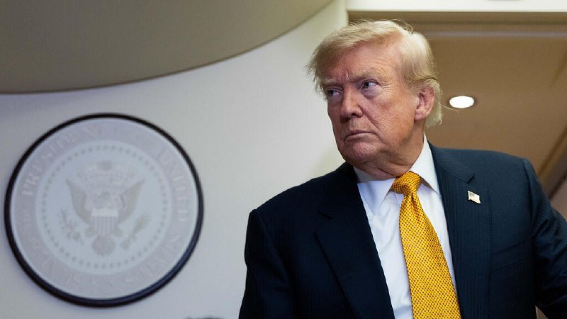 A man with blond hair wearing a dark suit and yellow tie stands beside the Presidential Seal of the United States.