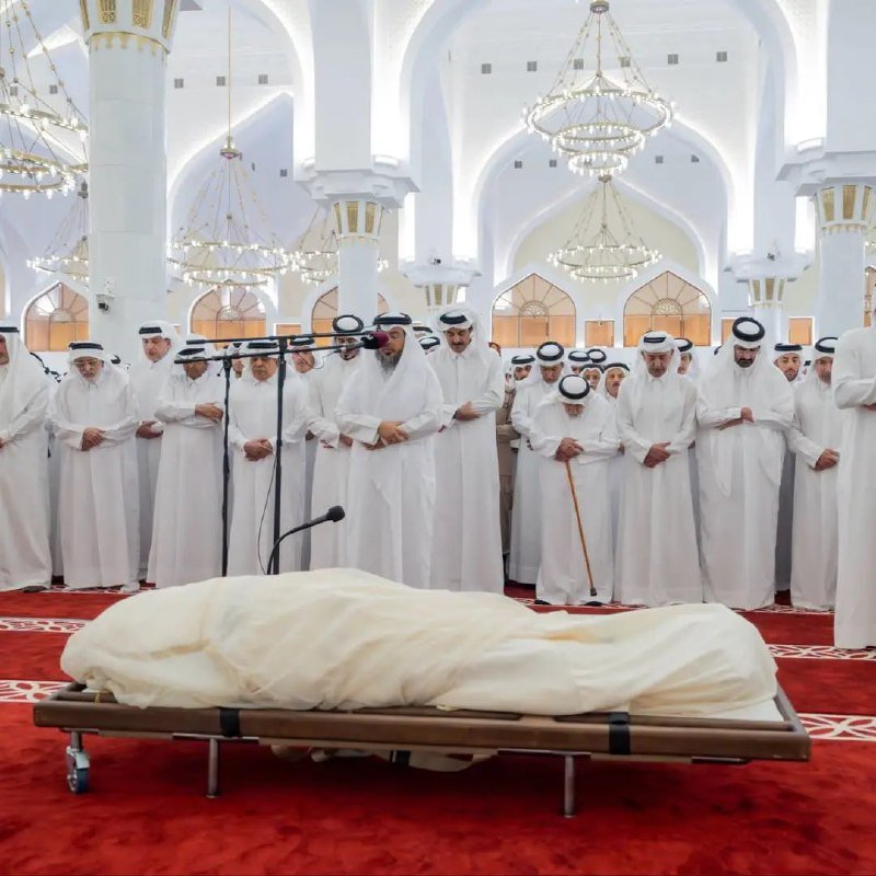 Men in traditional white robes and headdresses stand in prayer inside an ornate mosque with chandeliers, facing a body covered in white cloth on a wheeled bier atop a red carpet.