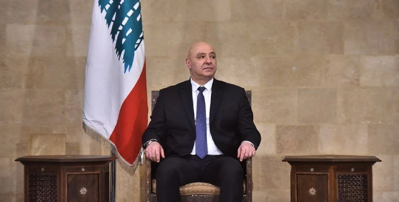 A bald man in a dark suit and blue tie sits in a wooden chair beside a Lebanese flag against a stone wall, flanked by small wooden tables.