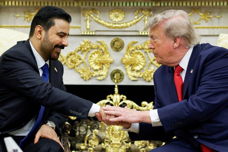 Two men in dark suits shake hands while seated in front of an ornate gold-decorated fireplace, both smiling.