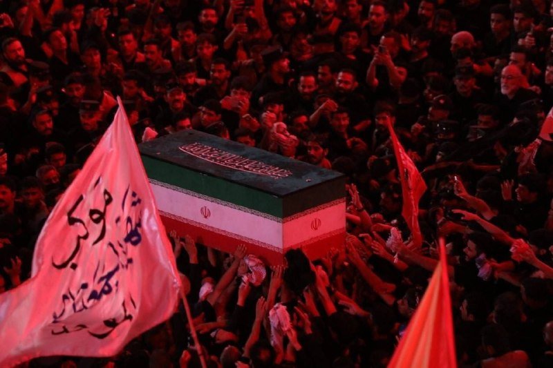 A flag-draped coffin is carried overhead by a dense crowd of mourners under red lighting, with white flags bearing Arabic script held aloft.