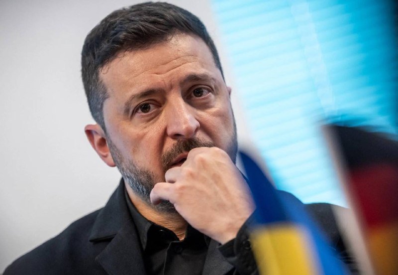 A man with short dark hair and a beard sits thoughtfully with his hand near his face, wearing a black shirt and jacket, with a blurred blue-and-yellow flag visible to the side.