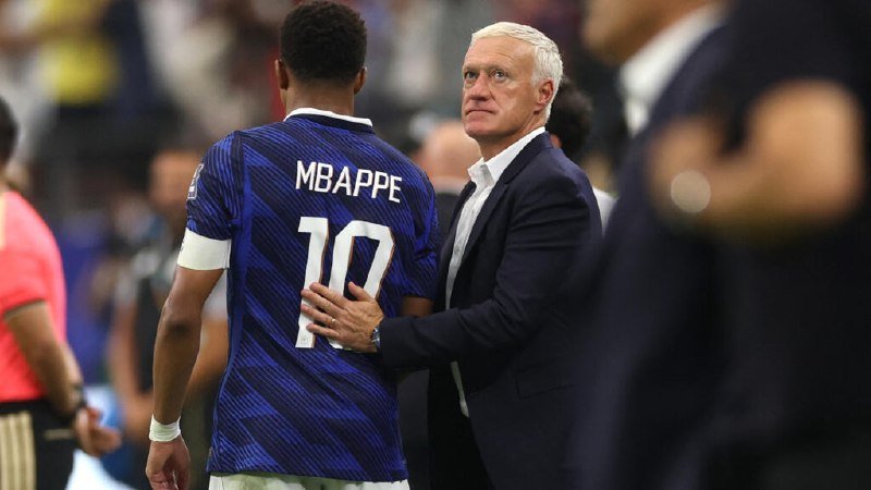 A man in a dark suit places his hand on the back of a soccer player wearing a blue jersey marked "MBAPPE 10," appearing to console or greet him on the field.