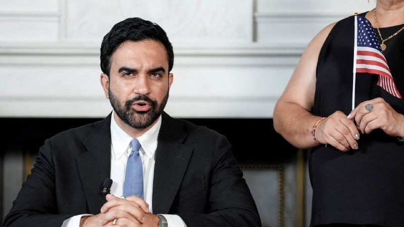A bearded man in a dark suit and blue tie speaks into a microphone, while a woman behind him in a black dress holds a small American flag.
