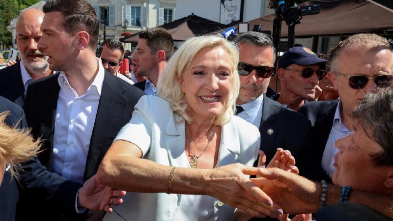A smiling blonde woman in a white jacket shakes hands while surrounded by a crowd of people, some in suits and sunglasses, outdoors near tents and a camera.