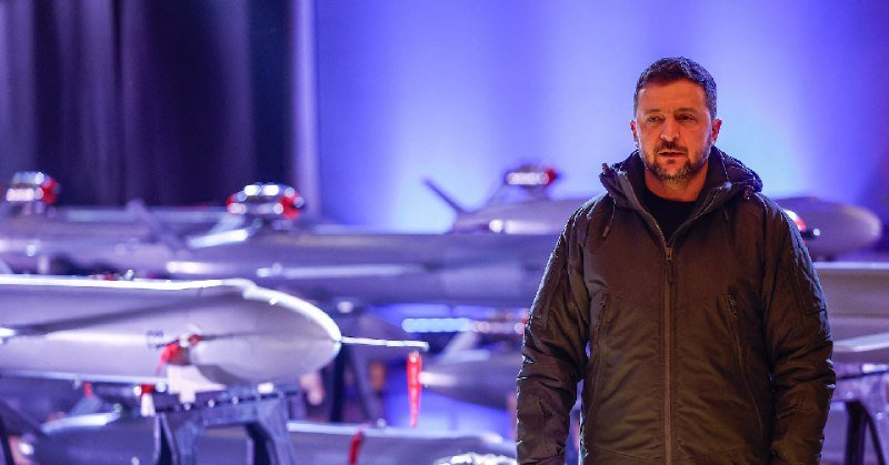 A man in a dark hooded jacket stands in front of several large drones displayed on stands under blue stage lighting.