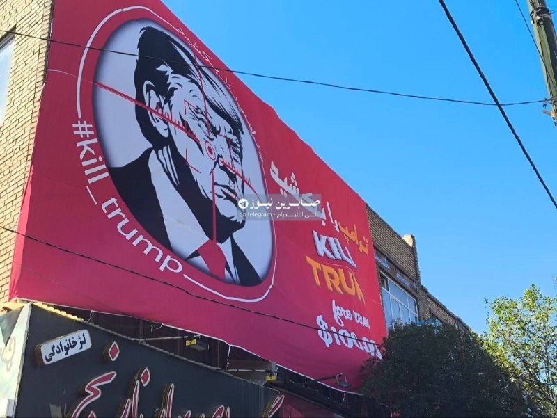A large red anti-Trump mural featuring a stylized portrait and the text "#KillTrump" is displayed on a brick building under a clear blue sky.