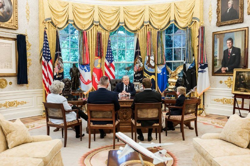 Five people sit around a desk in an ornate room with gold curtains, military flags, and a portrait, with a model airplane on the table.