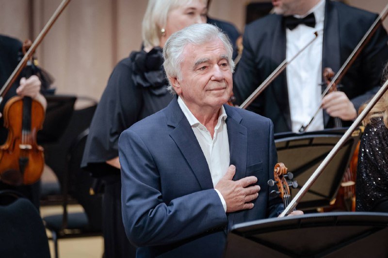 An older man in a navy blazer stands with his hand over his heart among orchestra members holding violins on a stage.