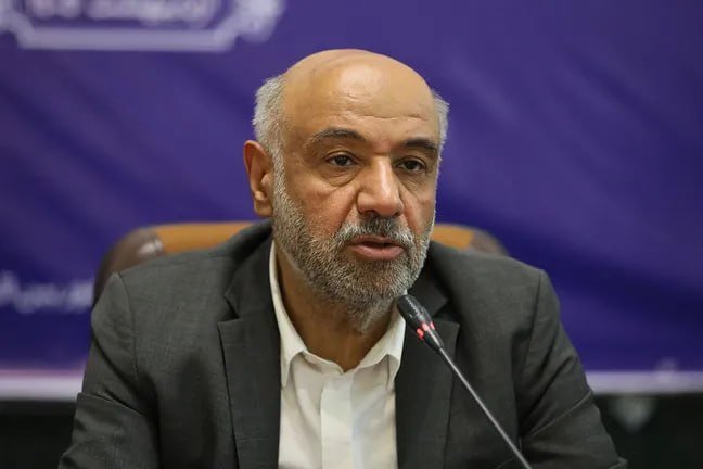 A bald, bearded man in a dark suit and white shirt speaks into a microphone against a purple backdrop with partial script visible.