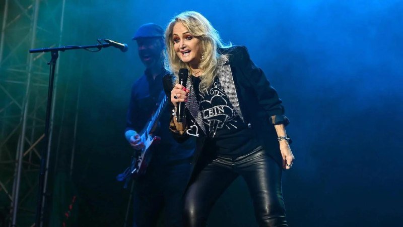 A blonde woman in a "Bonnie Tyler & A..." graphic t-shirt and black leather pants sings into a microphone on a blue-lit stage behind a guitarist.