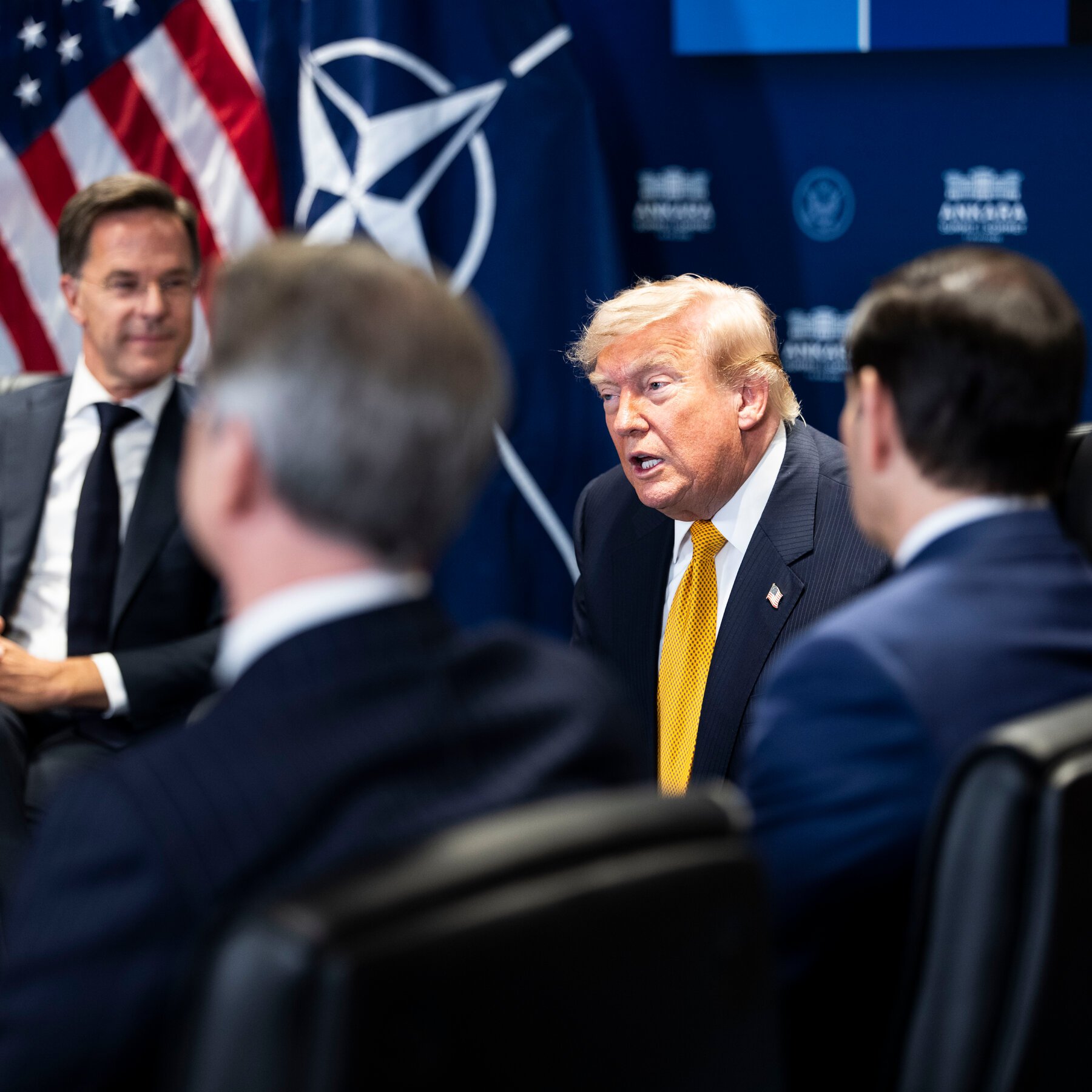 A blonde man in a dark suit and yellow tie speaks during a meeting, with an American flag and a NATO emblem visible on a blue backdrop behind him.