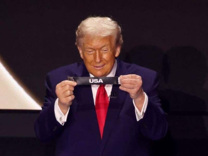 A man in a dark suit and red tie holds up a black wristband labeled "USA" while looking down at it against a dark background.