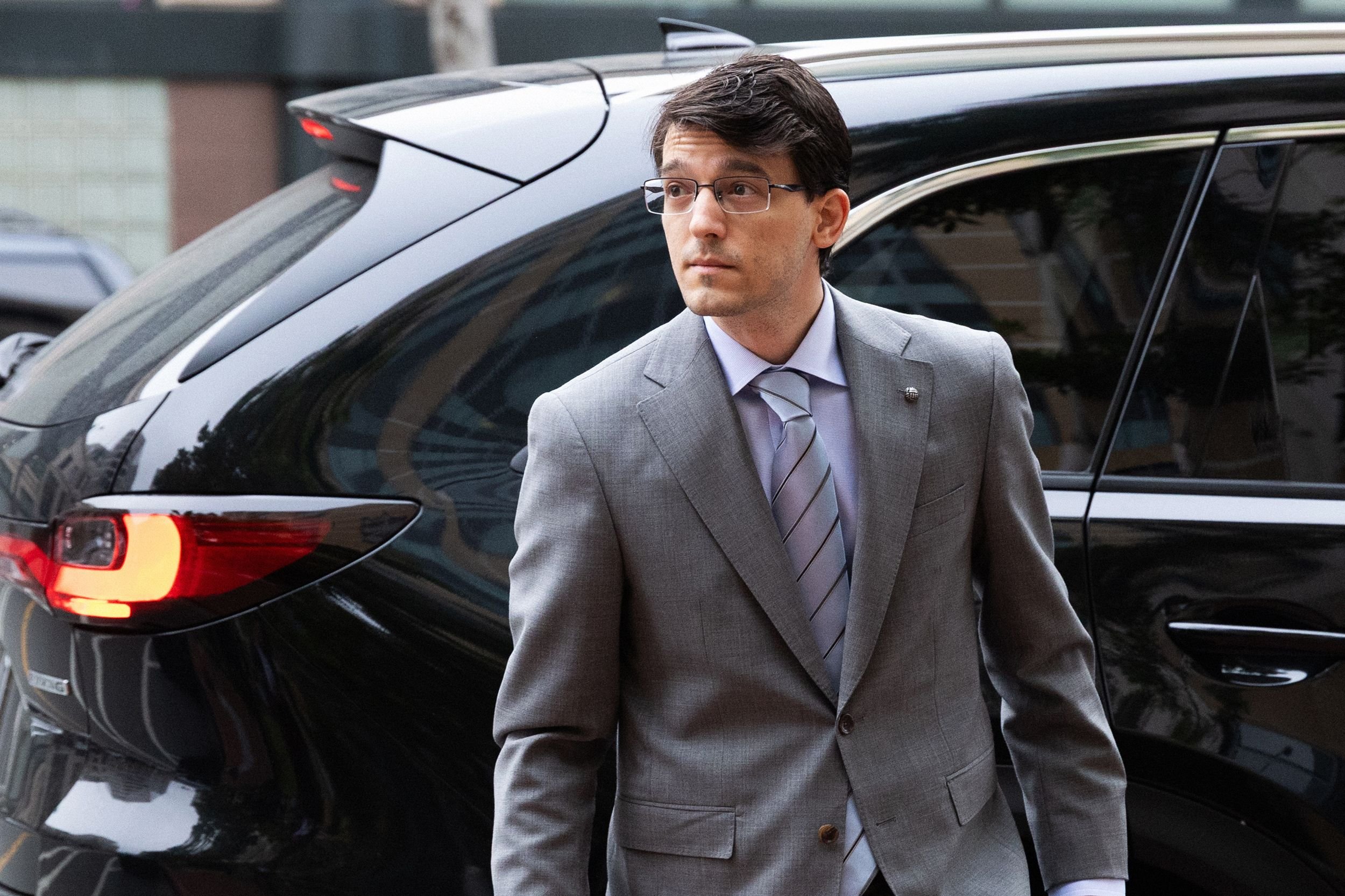 A bespectacled man in a gray suit and striped tie walks past a black SUV outside a building.