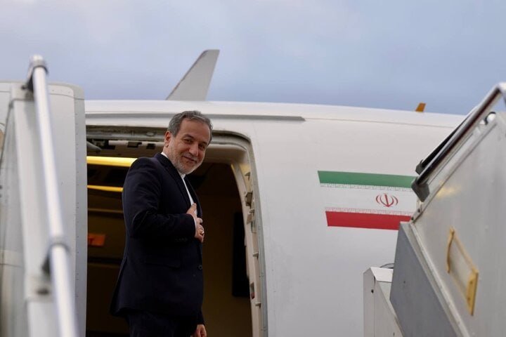 A bearded man in a dark suit stands in the doorway of an aircraft bearing the Iranian flag, descending the stairs at an airfield.