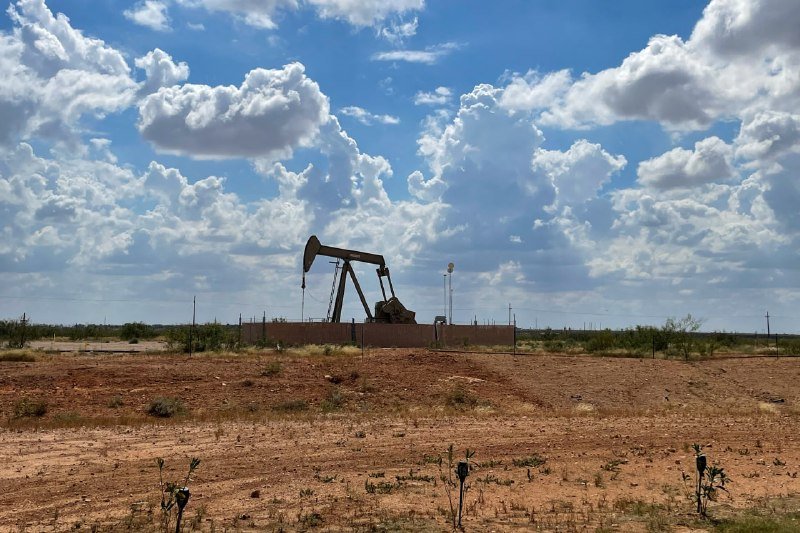A pumpjack operates in a fenced enclosure surrounded by sparse vegetation on reddish dirt under a partly cloudy blue sky.