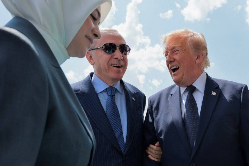 Two men in suits smile and laugh while speaking with a veiled woman against a partly cloudy sky, one man wearing dark sunglasses and an American flag pin.