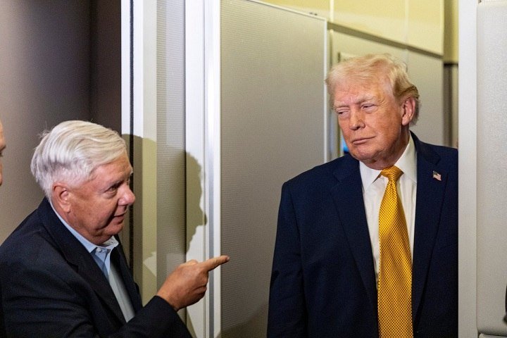 Two men in dark suits stand inside an aircraft cabin, one older with white hair pointing toward the other blonde man wearing a gold tie and an American flag lapel pin.