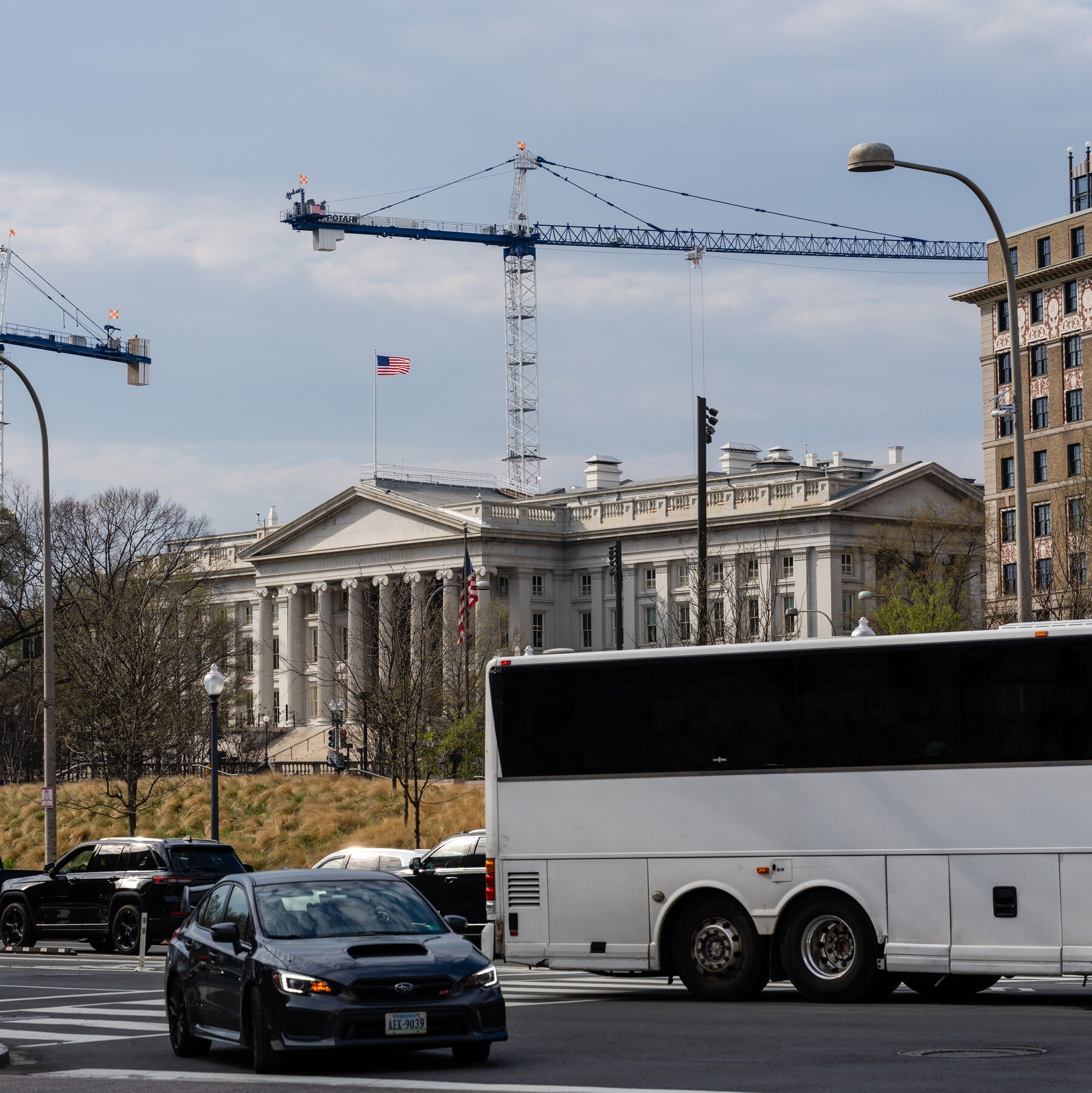 A dark Subaru WRX, a black SUV, and a white bus travel along a city street in front of a neoclassical white building flying the American flag, with a construction crane towering overhead.