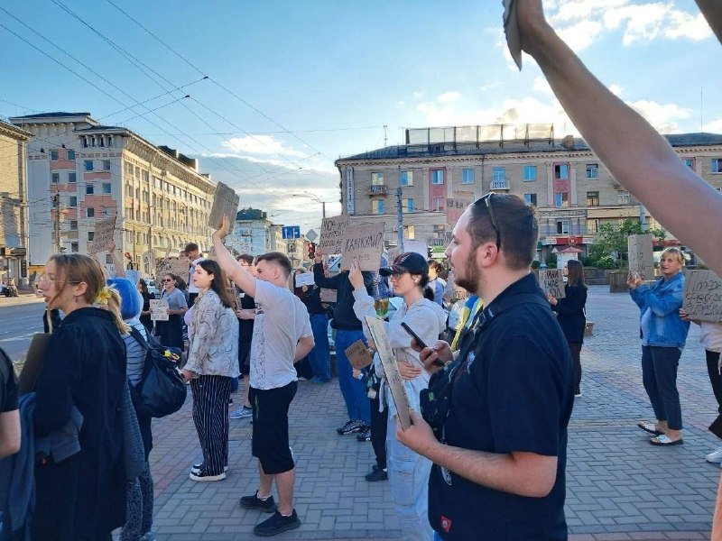 Crowd of people holding handmade signs gather in an urban plaza framed by multi-story buildings under a partly cloudy sky.