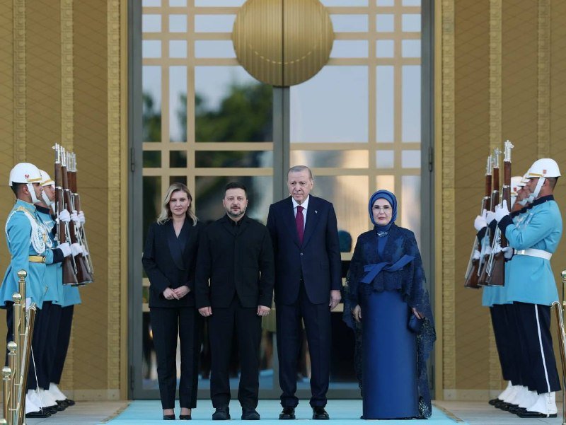 Four formally dressed individuals pose in front of ornate golden doors flanked by uniformed honor guards in light blue ceremonial attire holding rifles.