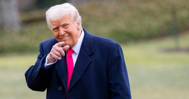 An older man with white hair, wearing a navy coat and red tie, smiles and points forward while standing on a lawn.