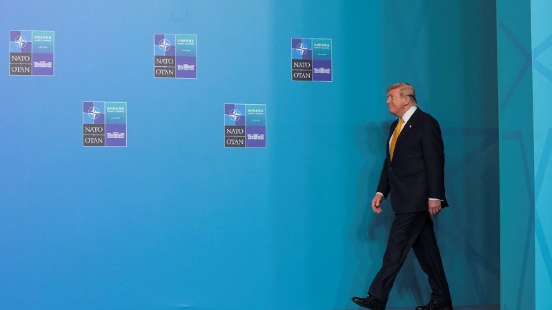 A man in a dark suit walks past a blue backdrop featuring NATO summit logos marked "ANKARA."