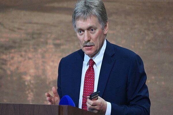 A gray-haired man with a mustache, wearing a dark suit and red tie, speaks at a podium while holding a mobile phone and a blue microphone.