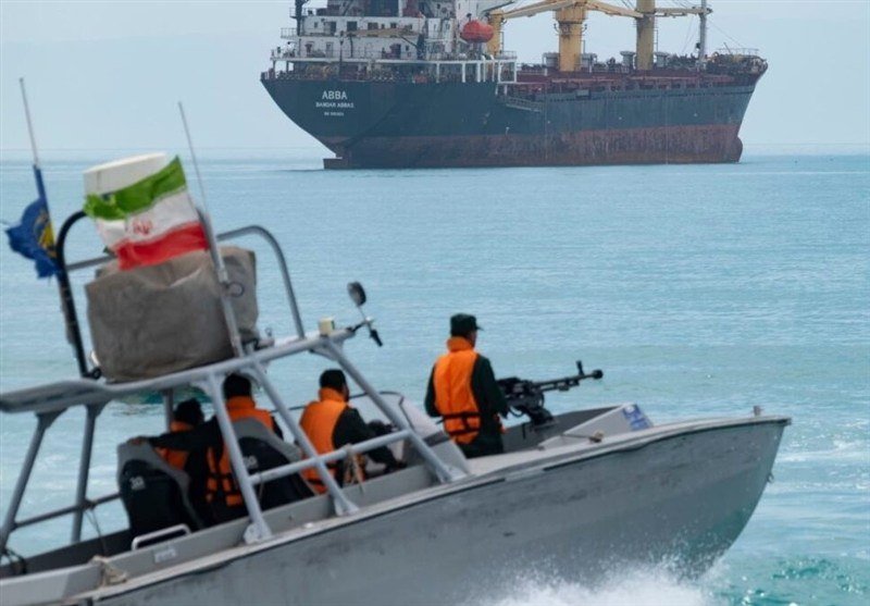 Armed personnel in orange life vests patrol from a small boat flying Iran's flag toward a large cargo ship labeled "ABBA" in open water.