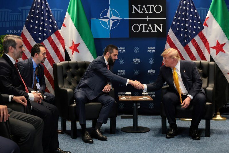 Two men in dark suits shake hands while seated in black armchairs, flanked by American and other flags with a "NATO/OTAN" backdrop.