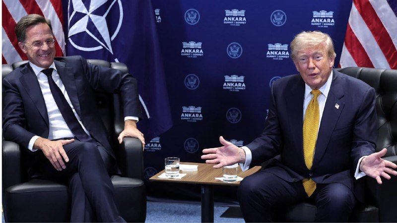 Two men in dark suits sit in black armchairs before a NATO flag and a backdrop reading "ANKARA SUMMIT," engaged in conversation with water glasses on a table between them.