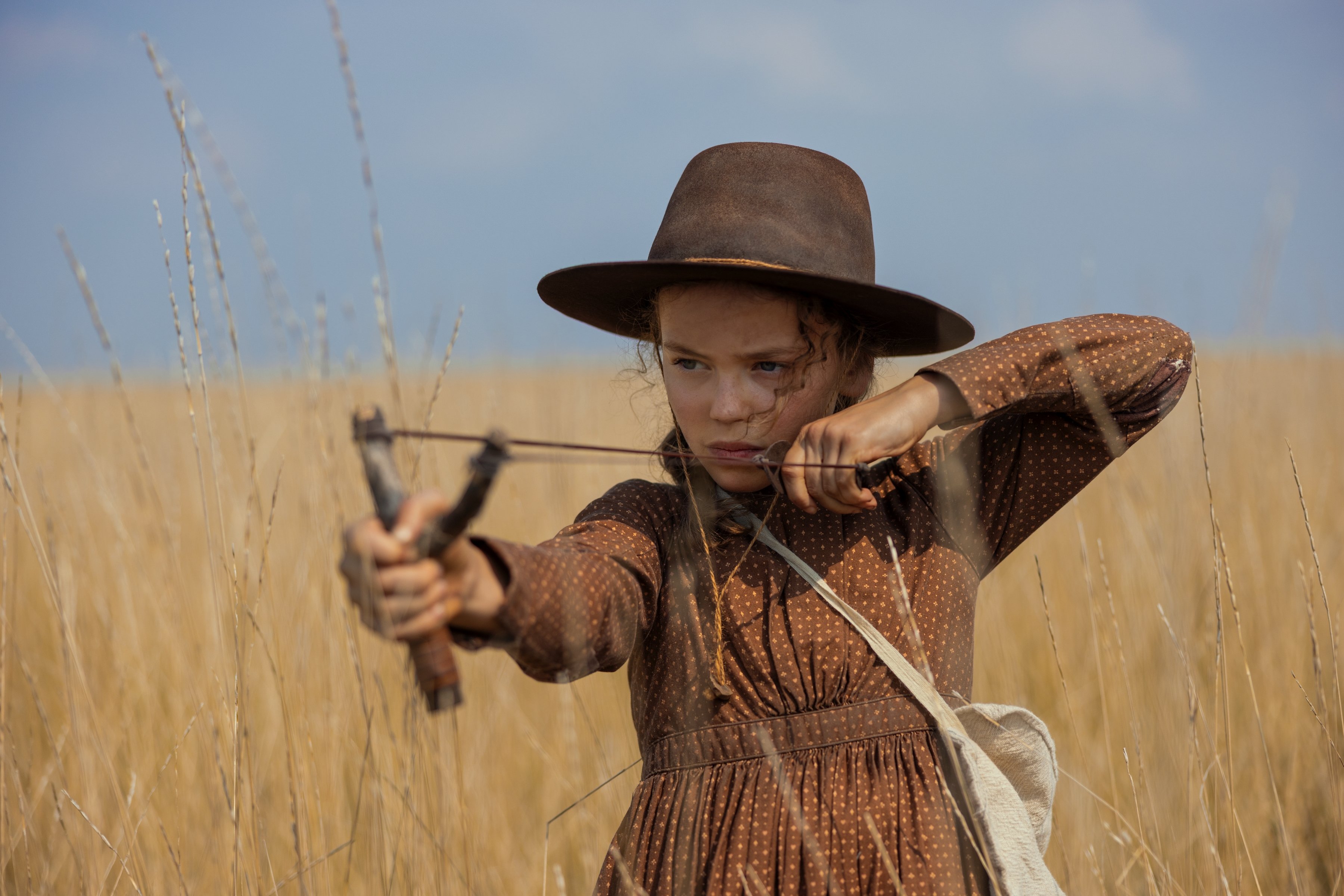 A young girl in a brown patterned dress and wide-brimmed hat aims a slingshot while standing in a field of tall, golden grass under a blue sky.