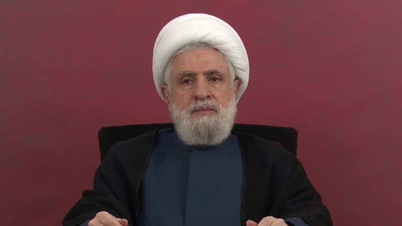 A bearded man wearing a white turban and dark religious robes sits against a solid maroon background.