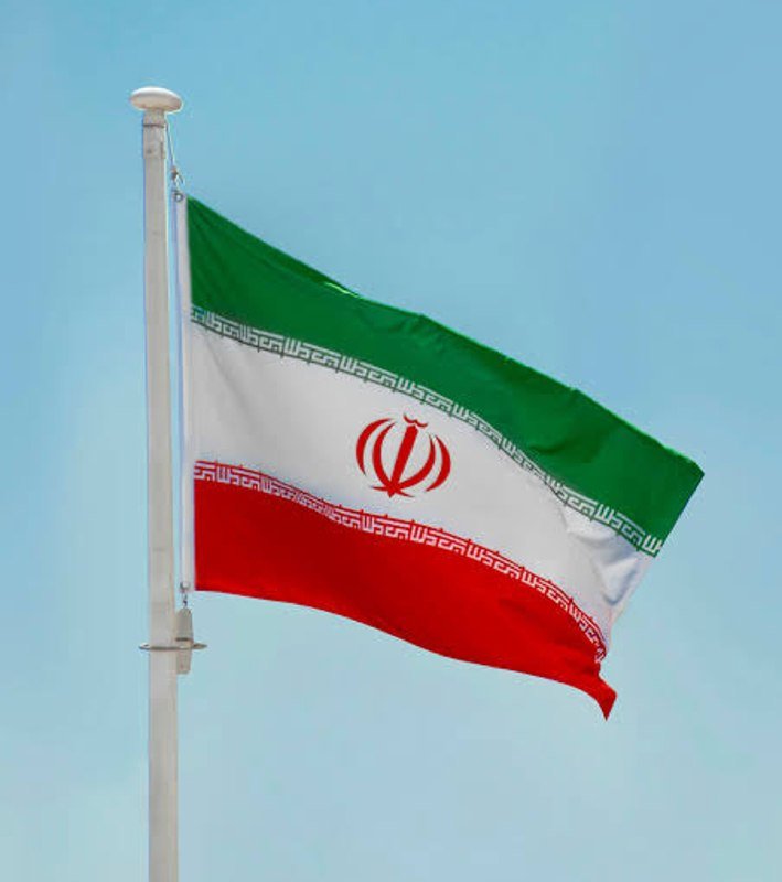 The flag of Iran, featuring green, white, and red horizontal stripes with a central red emblem, waves on a flagpole against a clear blue sky.