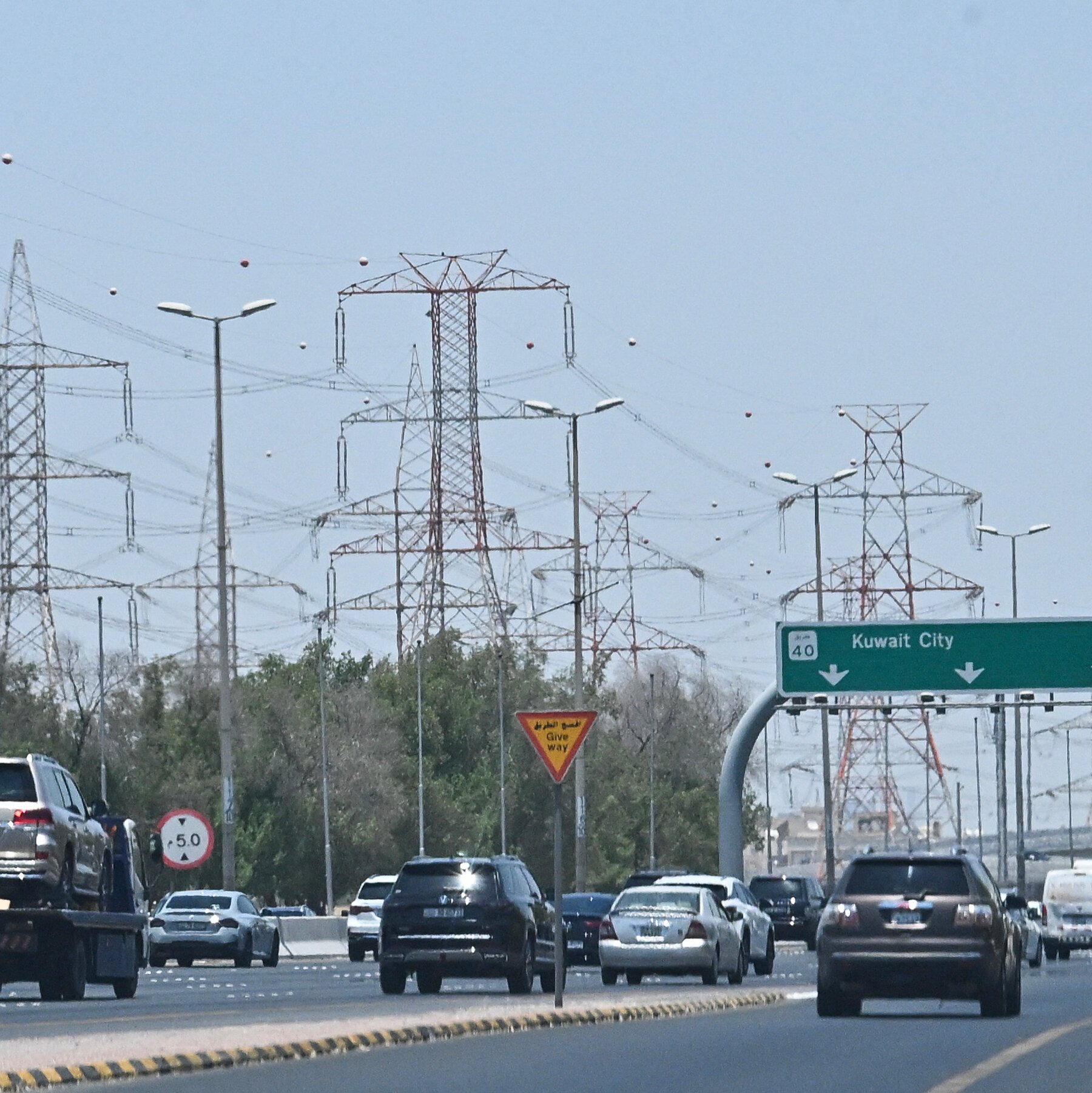 Vehicles travel along a multi-lane highway beneath tall electrical transmission towers, with a green overhead sign reading "Kuait City."