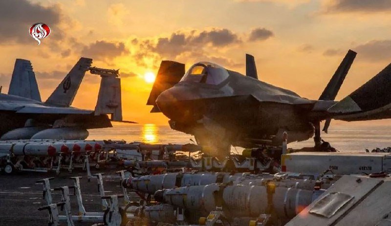 Military aircraft and rows of mounted bombs sit on a deck at sunset, with the ocean visible in the background.