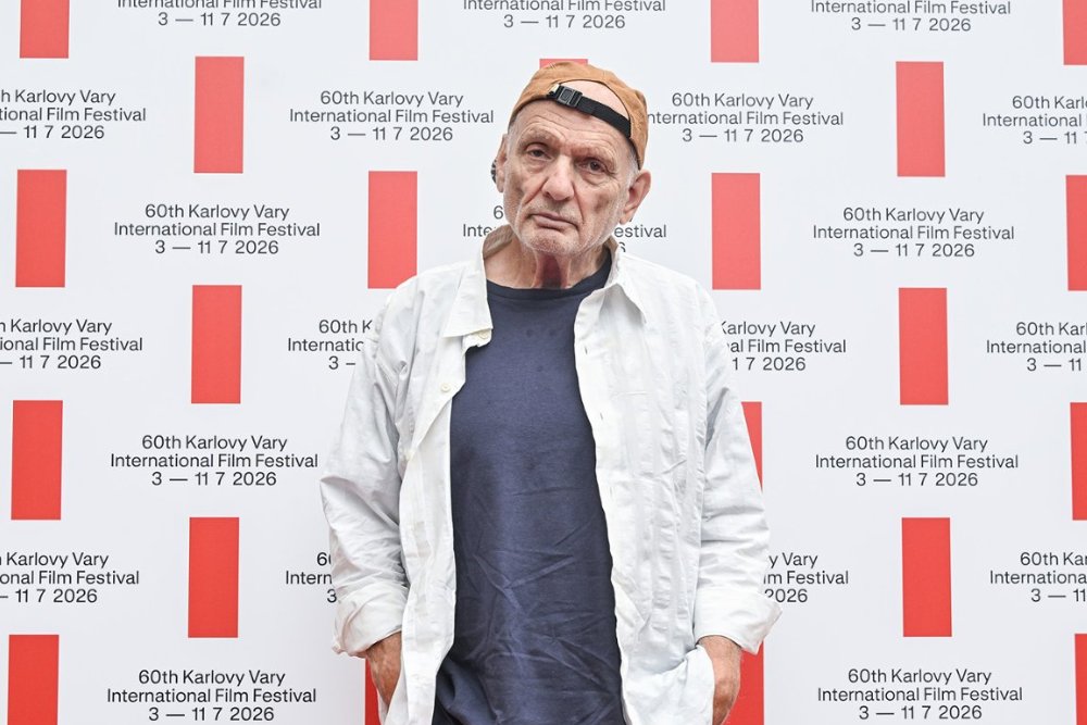 A man wearing a backward tan cap, unbuttoned white shirt, and navy t-shirt stands against a backdrop repeatedly printed with "60th Karlovy Vary International Film Festival 3 — 11 7 2026."