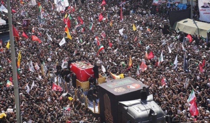 A massive crowd waves colorful flags during a procession carrying a red-draped casket on a decorated truck through a city street.