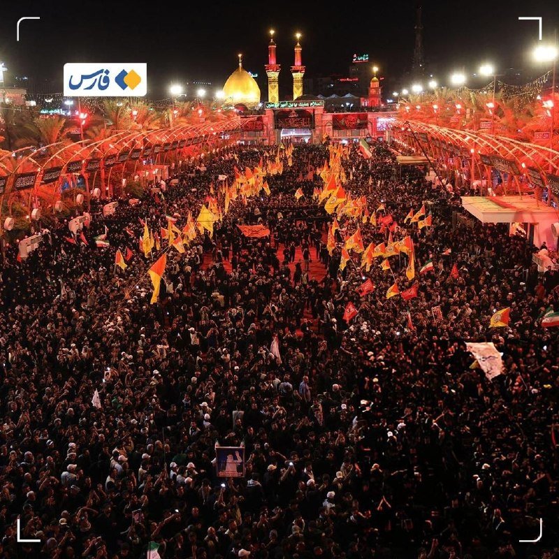 Large nighttime gathering of people holding yellow and red flags between arched canopies, gathered before a golden-domed shrine illuminated in the background.