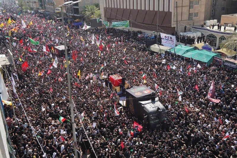 A massive crowd fills a city street, surrounding an elevated black-draped casket as participants wave red, black, and multicolored flags during a procession.