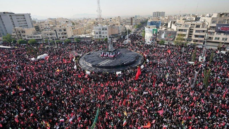 A massive crowd holding red and green flags fills a city square, surrounding a central stage with banners and a large portrait, as people gather in the surrounding streets.