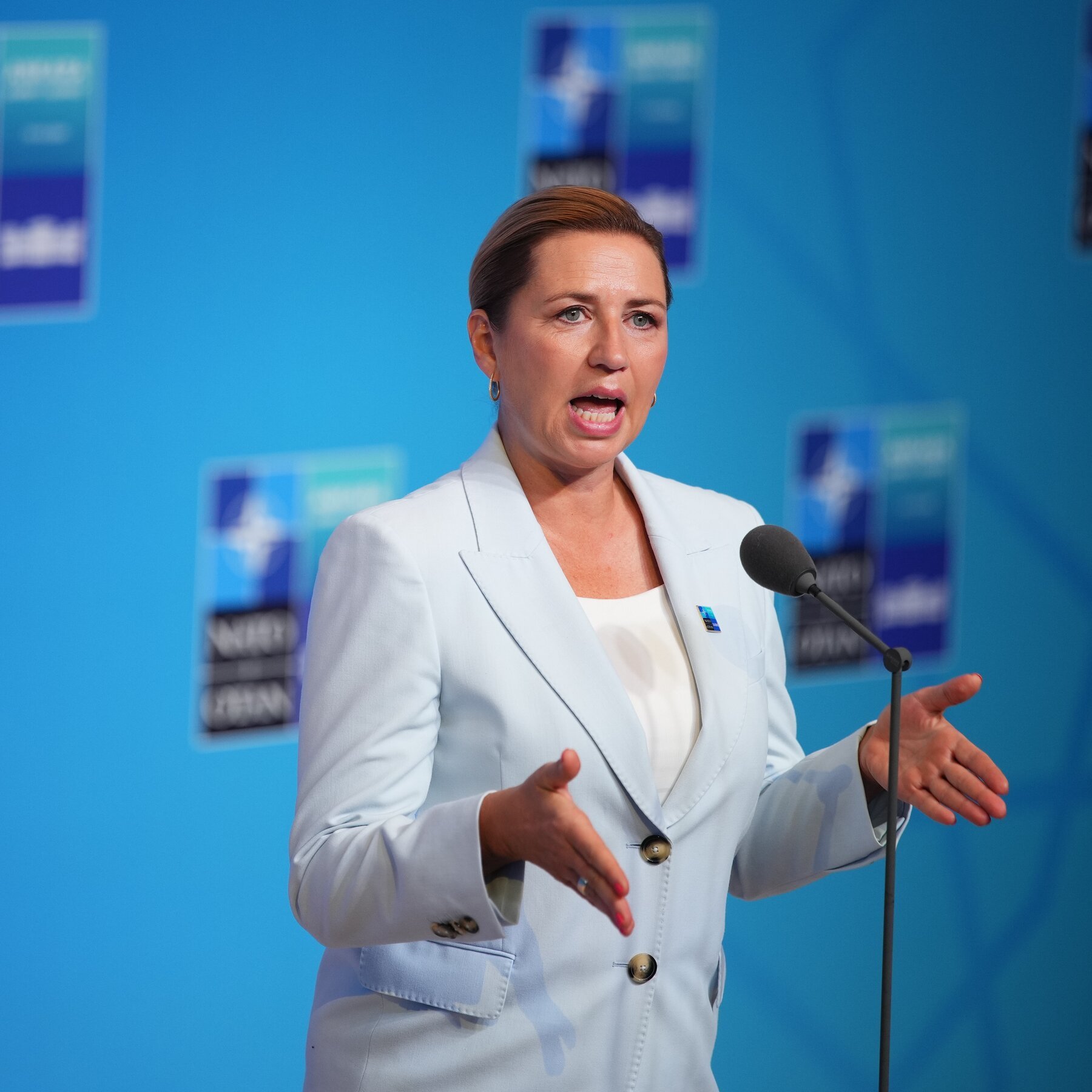 A woman in a light blue blazer speaks at a microphone, gesturing with both hands against a blue backdrop featuring repeated graphic logos.