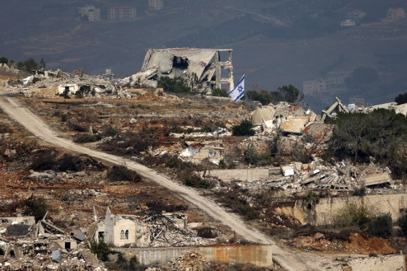 A destroyed hillside settlement with rubble, a partially collapsed concrete building, and an Israeli flag flies amid scattered debris and a dirt path winding through the ruins.