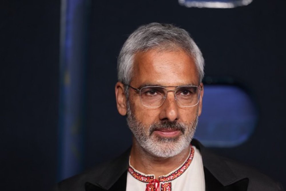 A gray-haired man with a beard and glasses wears a black blazer over an embroidered white shirt, standing against a dark backdrop.
