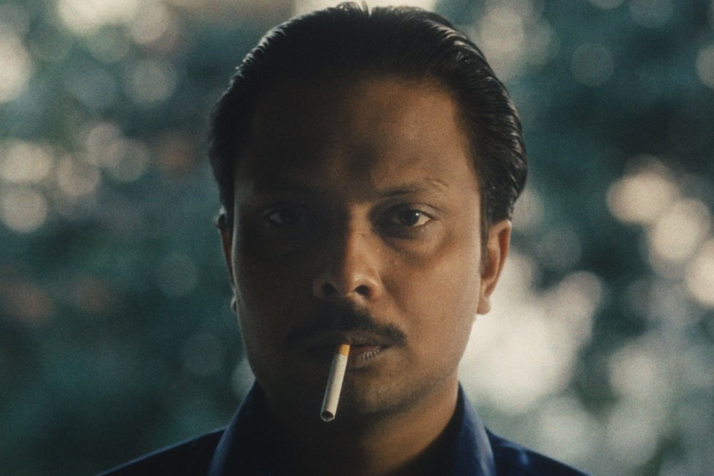 A man with slicked-back dark hair and a mustache holds an unlit cigarette between his lips, gazing off-camera against a blurred bokeh background.