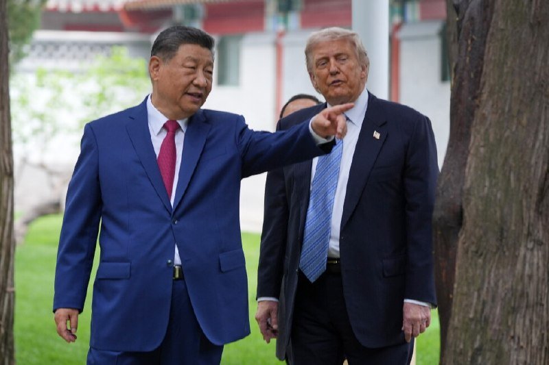 Two men in business suits walk outdoors near trees and a building, one gesturing with his arm extended while the other looks on.