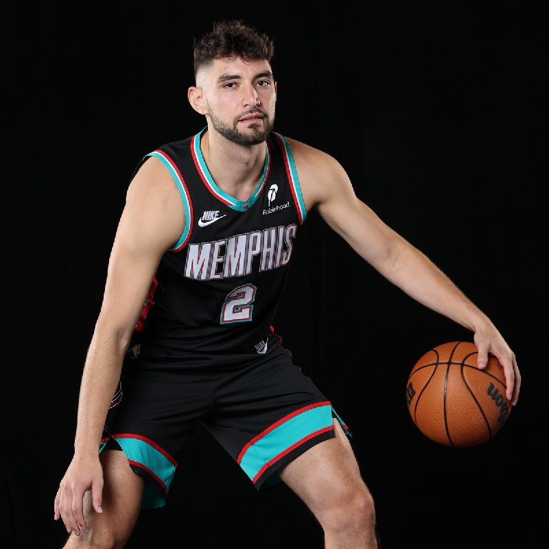 A bearded basketball player wearing a black Memphis jersey with the number 2 poses against a black backdrop while holding a basketball in his left hand.