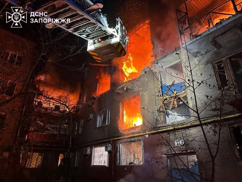 A multi-story residential building is engulfed in nighttime flames, with a firefighter in an extended aerial ladder spraying water onto the burning structure.